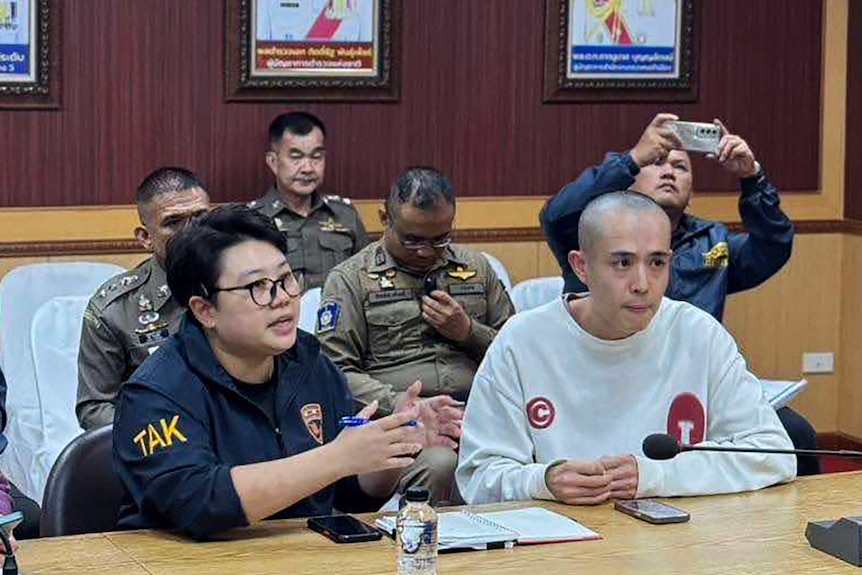 Wang Xing sitting in a white jumper at a wooden desk alongside members of the Thai police in dark and green jumpsuits