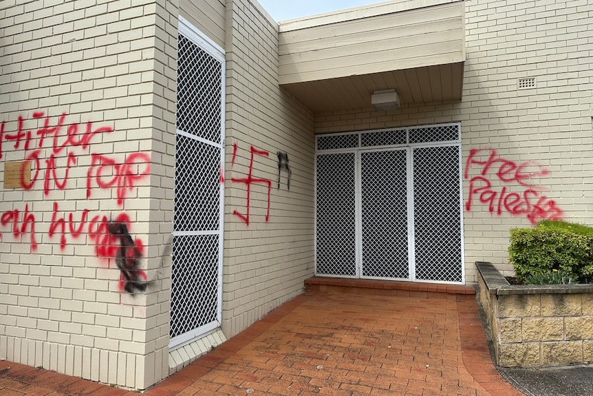 Anti-Semitic graffiti mentioning Hitler and showing a swastika along a brick wall.