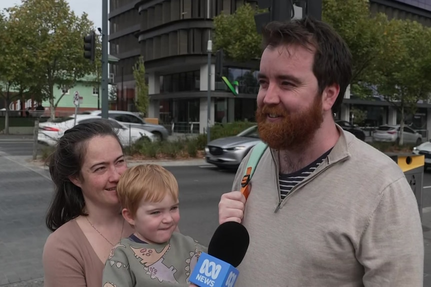 A woman holds a young boy with light red hair and looks at a man with brown hair and a reddish-brown beard.