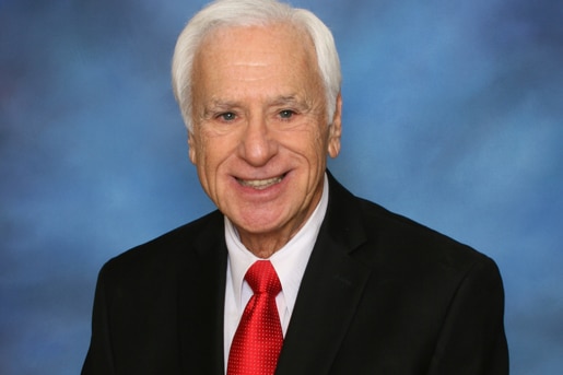 Harvey Kaye smiles, wearing black suit jacket, white shirt and red tie.