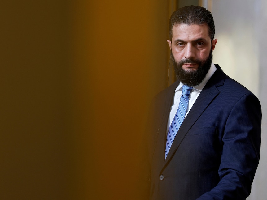 A man in a blue suit, blue tie and white shirt looks sternly at something out of frame