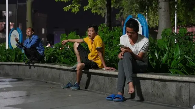 三名失业男子坐在公园的窗台上，彼此之间保持着一定的距离。一个穿着蓝色衬衫，蹲着看手机；另一位身穿亮黄色衬衫的人用手撑着身体，一只脚踩在窗台上，看着镜头；他旁边是另一个穿着白衬衫的男人，翘着腿，低头看着手机。