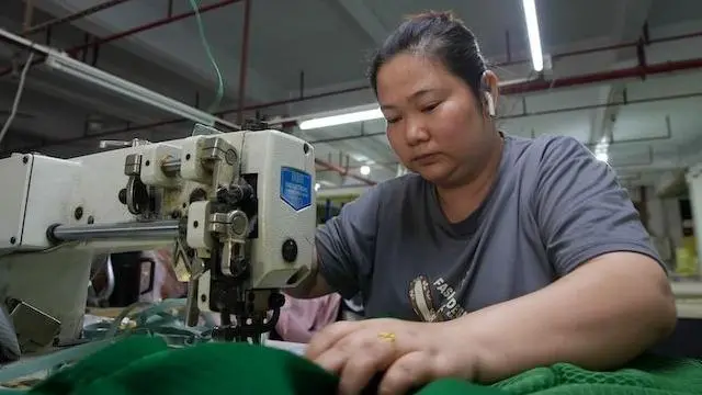 沙发工厂里，一位身穿灰色T恤的女士坐在缝纫机前。她在桌边戴着白色耳机，手里拿着一大块绿色布料。