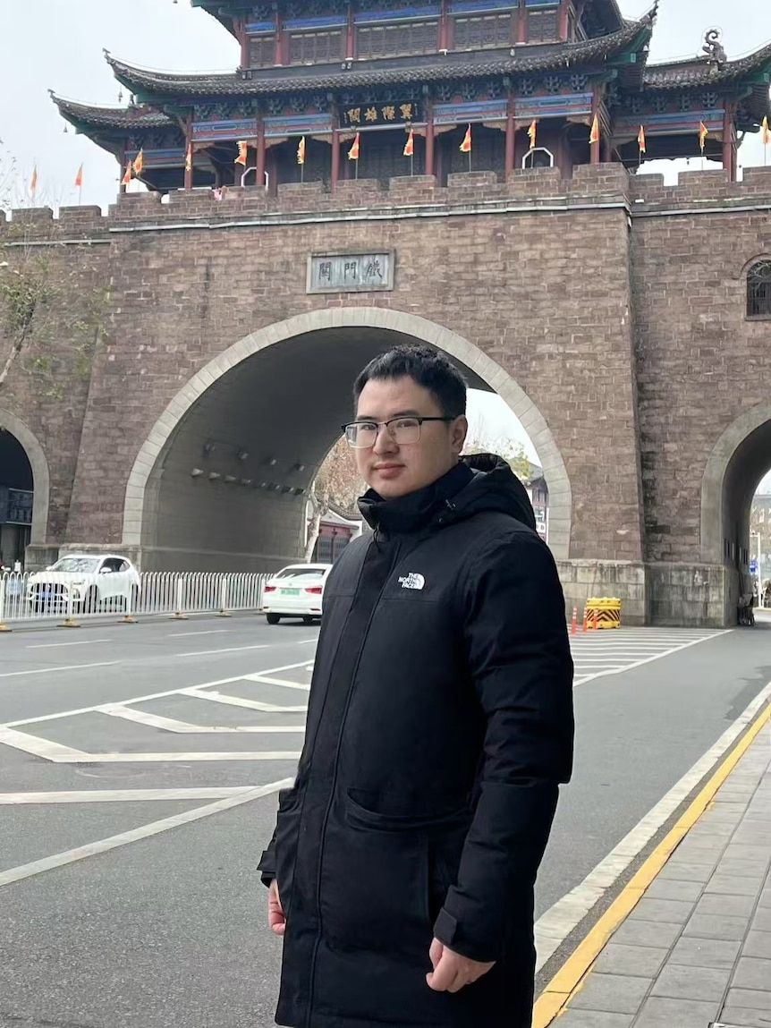 A man in a black coat stands in front of a Chinese-style city gate.