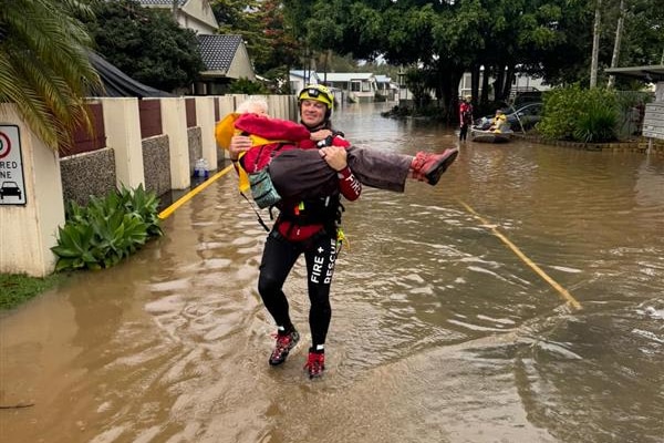 Man in emergency services uniform, wearing yellow helmet carries an elderly woman through floodwaters