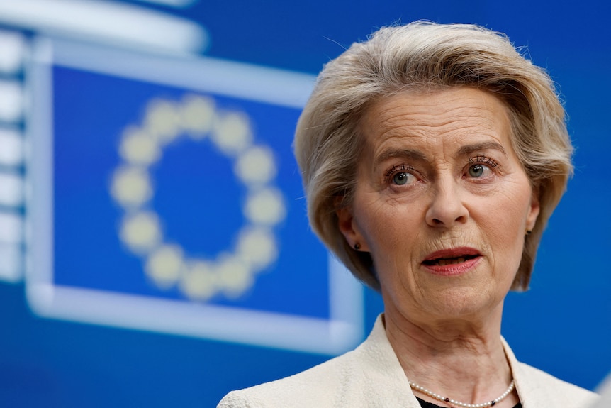 EU president wears speaking in front of a blue EU flag at a conference 