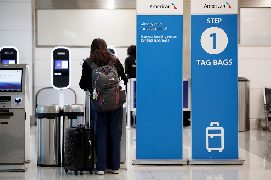 A female traveller in black clothing with a black bag next to a blue and white American Airlines bag tag and drop airport site