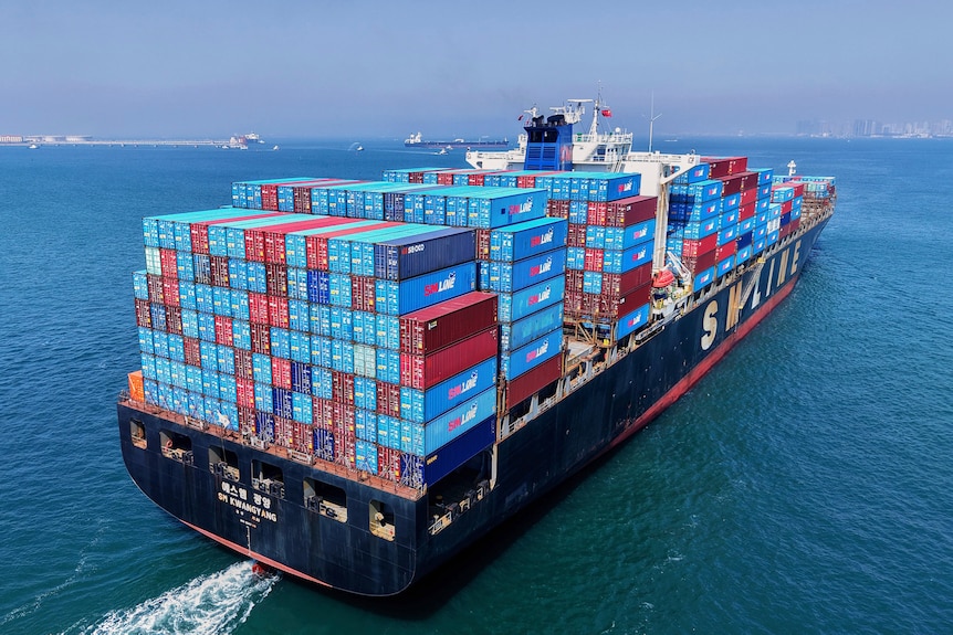 A loaded container ship sails off from a container terminal across calm blue waters.