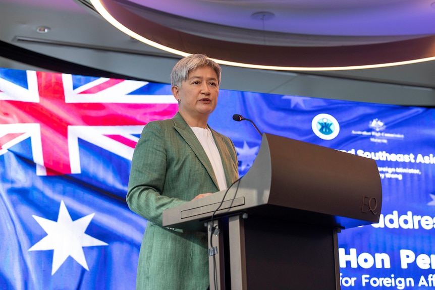 Penny Wong stands at a lectern with an image of an Australian flag in the background.