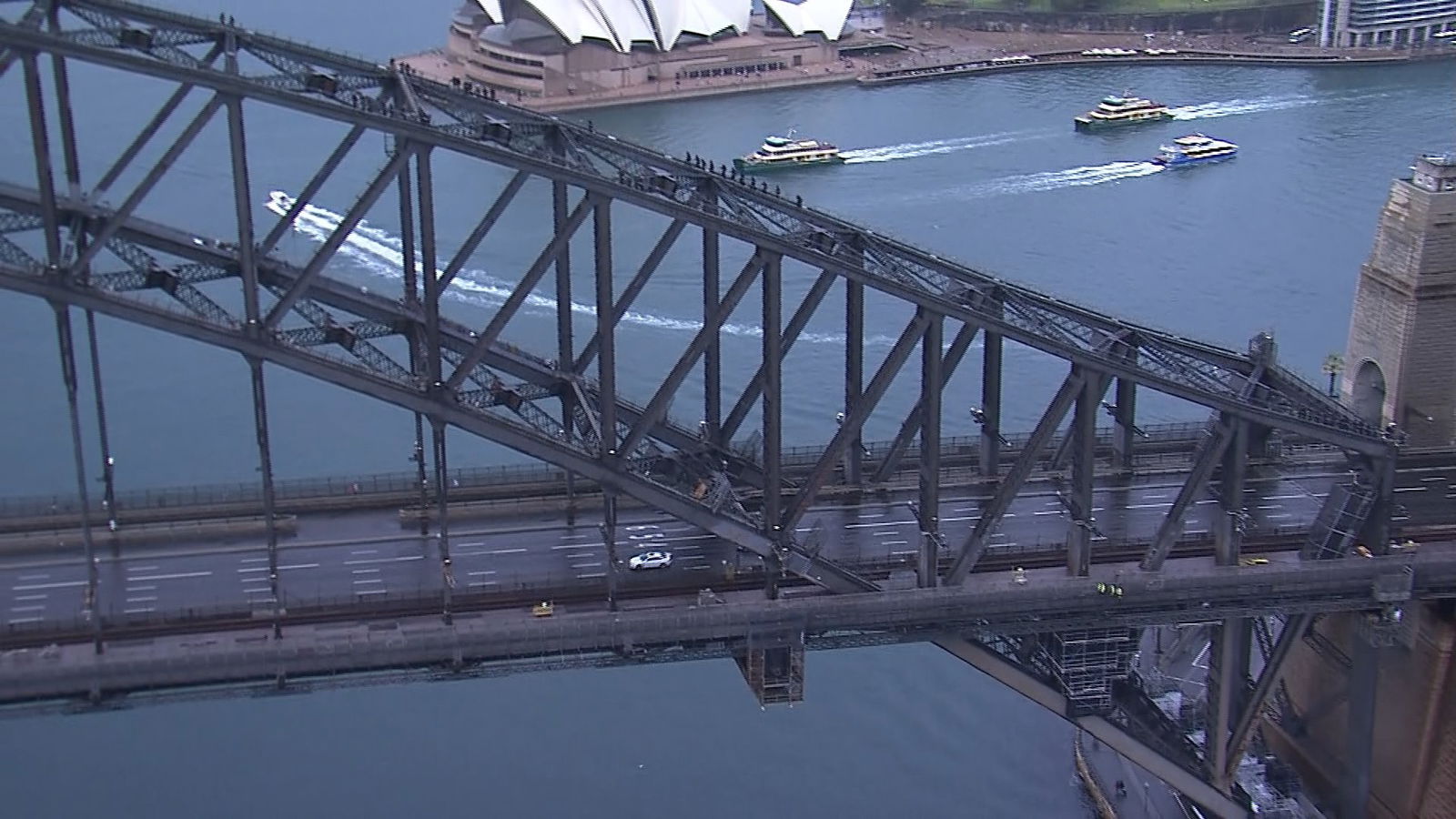 One white car on Sydney Harbour Bridge from the air. Ferries in water near Sydney Opera House in the background.