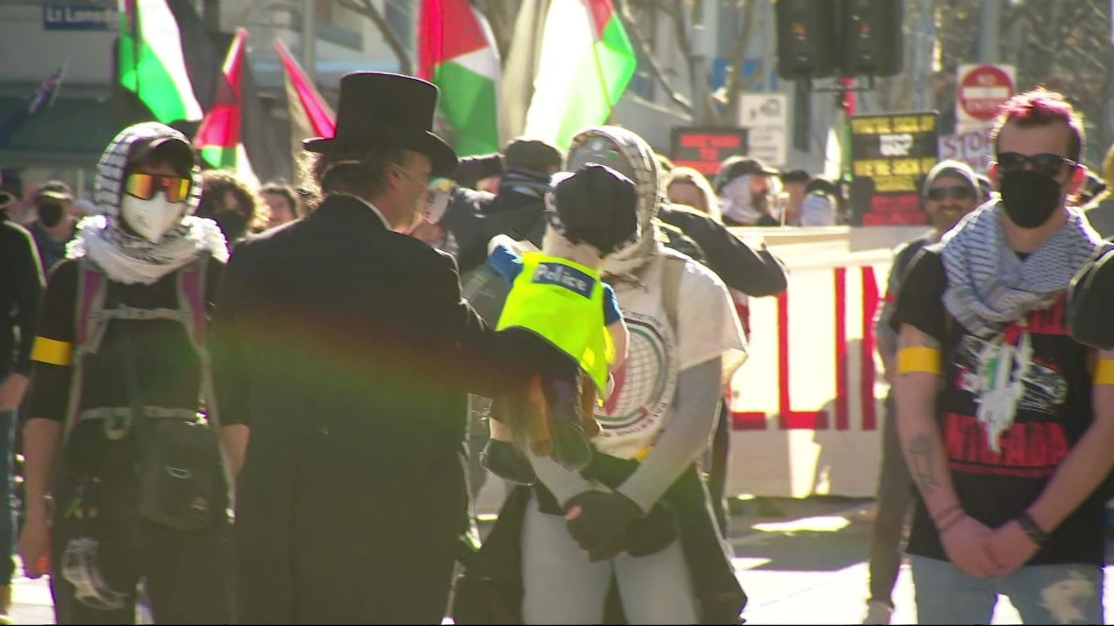 一名身穿黑衣、头戴黑色高顶礼帽的男子手持警察人偶,与手持旗帜的蒙面人群站在一起。