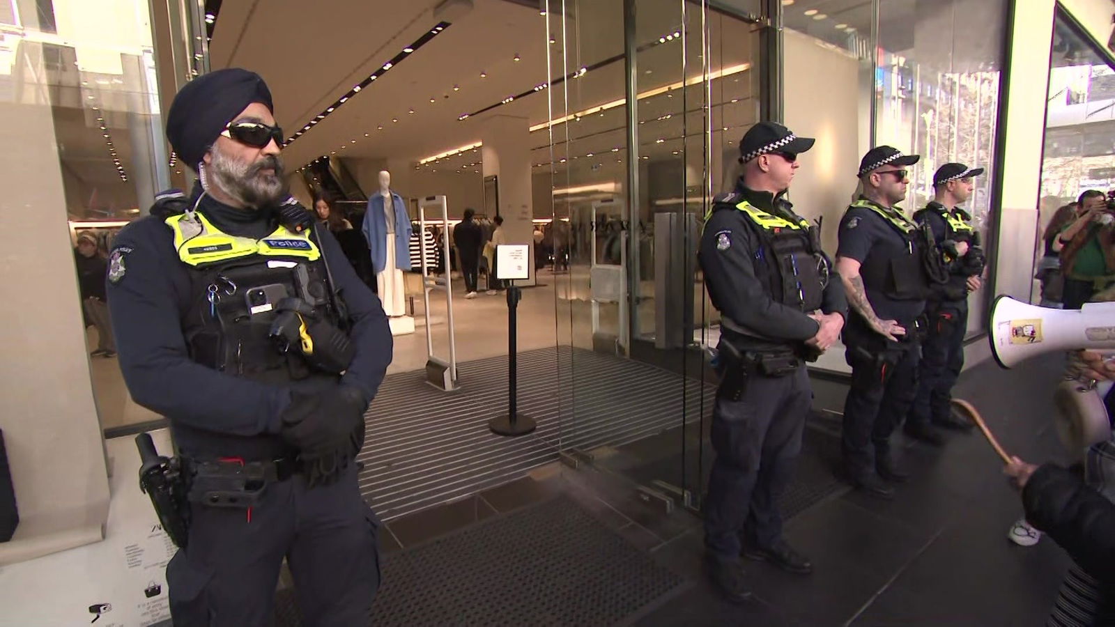 四名身穿海军制服和黄色背心的警察站在商店的玻璃墙边。