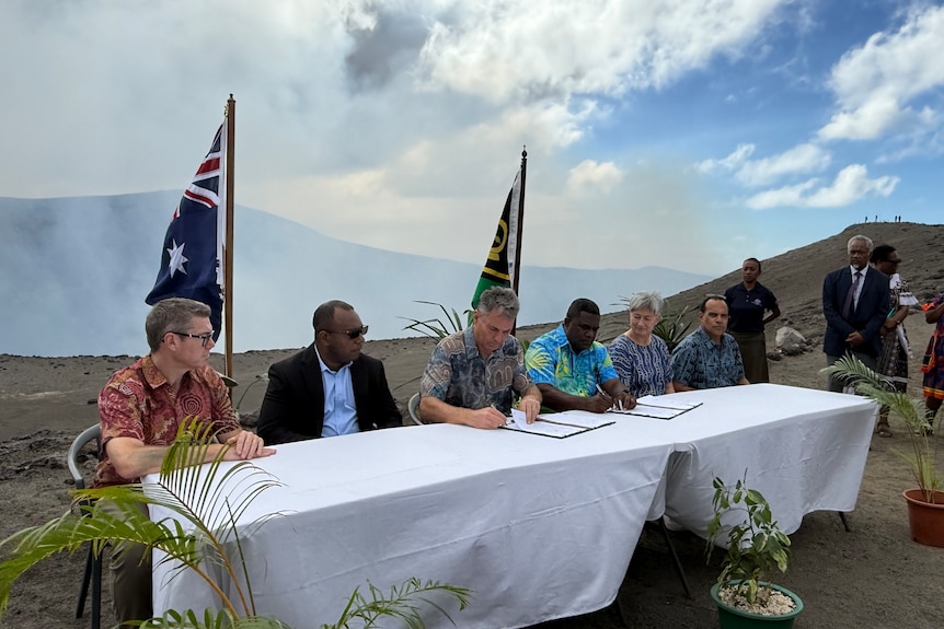 Six people sit behind a table signing a document with smoke rising from a crater behind them