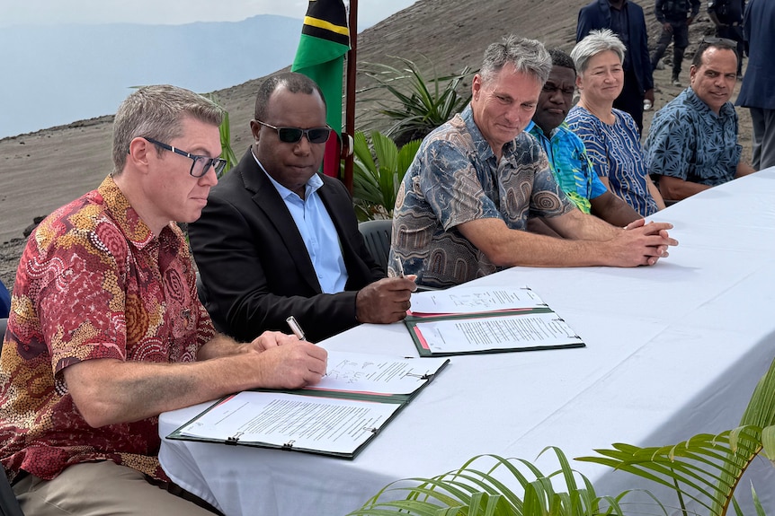 Six people sit behind a white long table looking at documents being signed