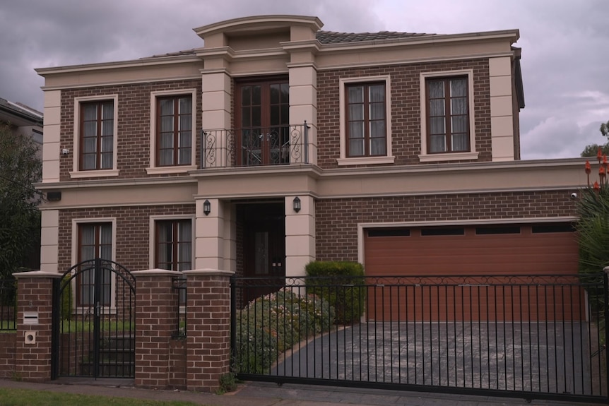 A double storey brown and cream brick home behind a brown brick fence with a black wrought iron gate on a cloudy day.