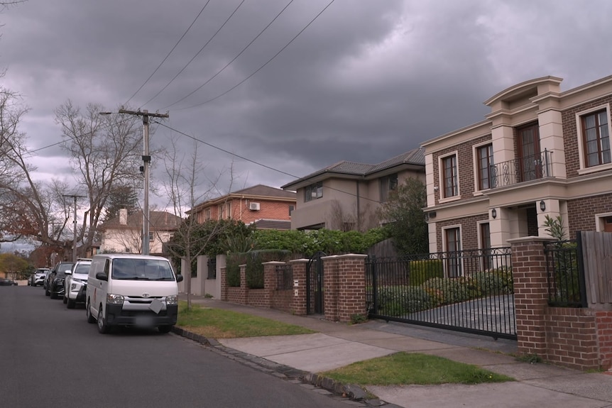 A two storey brown and cream brick home on a street lined with parked cars on a cloudy day.