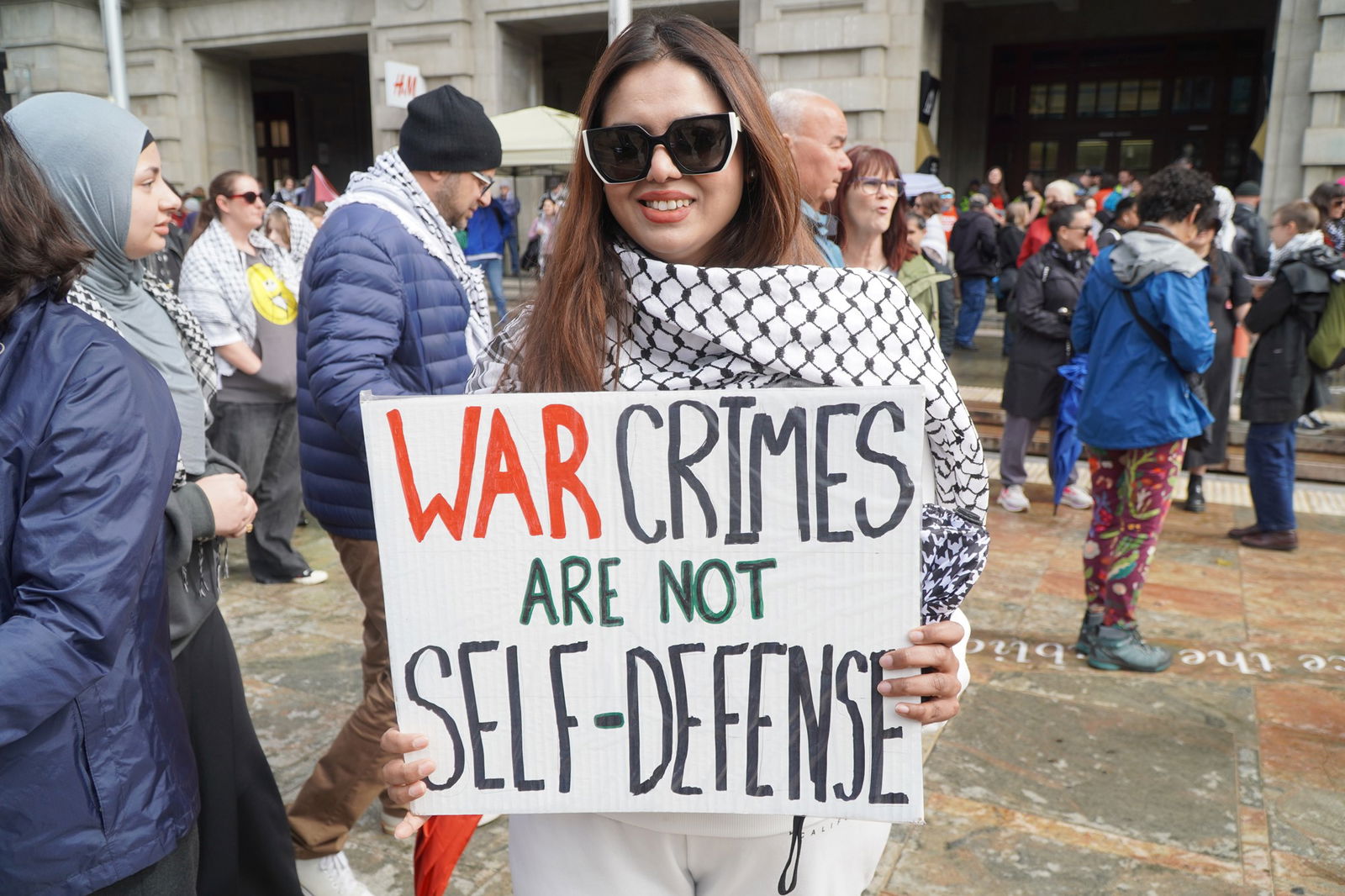 一名戴着大墨镜、肩上披着头巾的妇女举着一块牌子，上面写着：“战争罪不是自卫。”