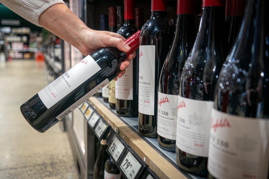 A hand removes a bottle of Penfolds from a shelf.