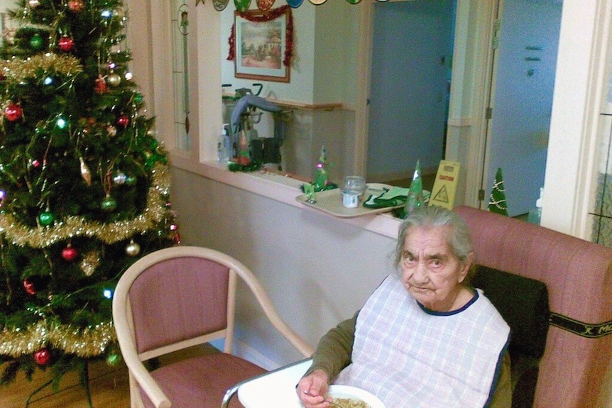 An elderly woman sitting with Christams tree and decorations around her.