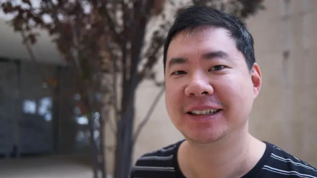 一名留着短黑发的男子站在建筑物和一棵树前，对着镜头微笑