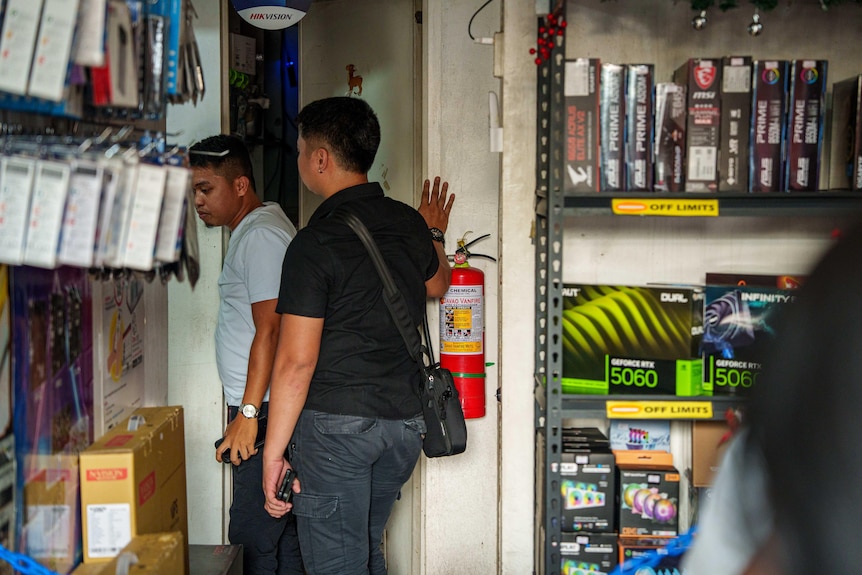 A man wearing a black tshirt stands with one palm up against the wall. Another man wearing stands in front