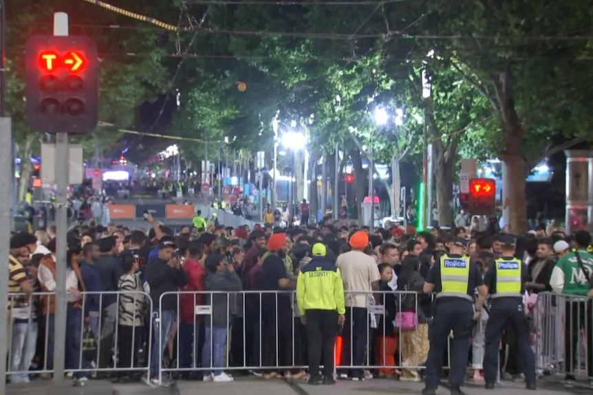 Police in yellow high vis vests stand in front a large crowd which is behind temporary fencing on a city street at night.
