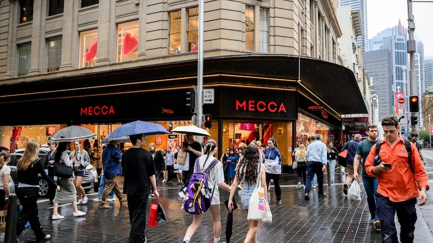 A crowd of people carrying umbrellas and shopping bags walk along a street on a rainy day.