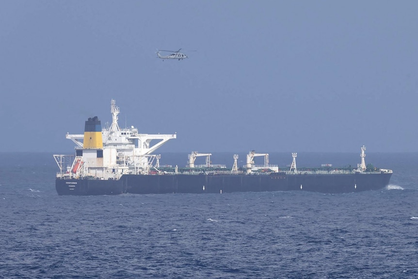 A US military helicopter flying over a large oil tanker at sea.