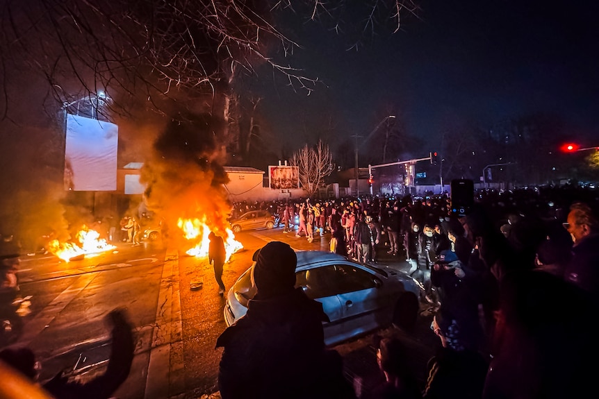 A group of protesters on a street at night with fires burning on the road.