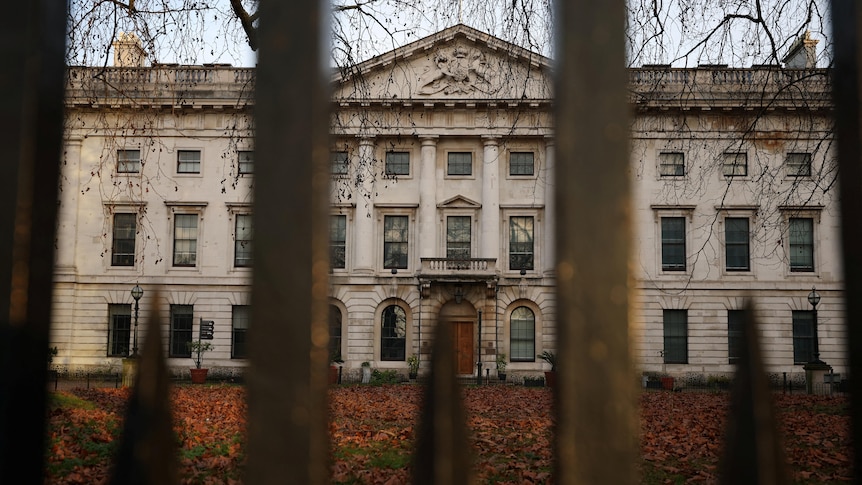A multi-storey building sits behind iron gates with brown leaves sitting on the lawn in front of it