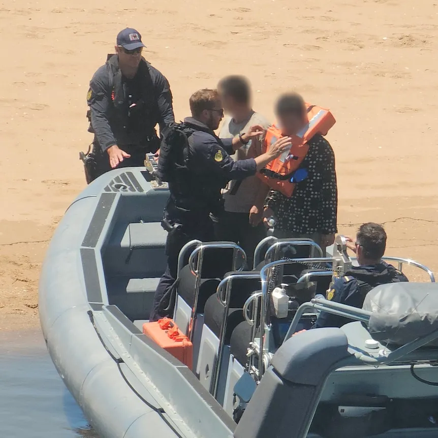 Men in uniiform hand lifejackets to other men on a boat moored on orange sand.