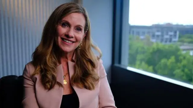 英曼·格兰特穿着浅粉色外套和黑色衬衫，微笑着看着镜头。她坐在窗边。