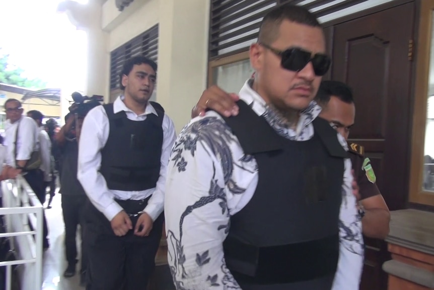 Two men wearing collared white shirts and vests walk into court with their hands tied.