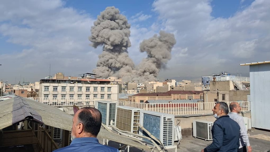 Three men watch from a building rooftop as smoke rises on the skyline of a city.