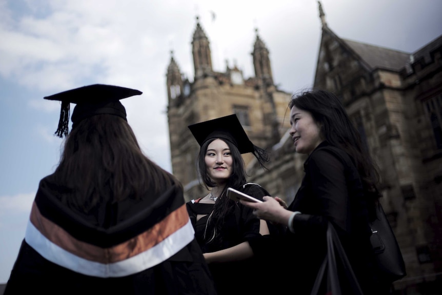 A trio of female international graduates stand outside a university building in academic gowns.