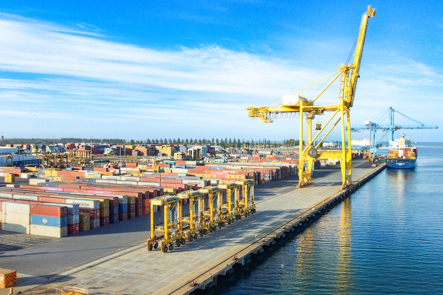 A wide shot of a port, with stacked shipping containers, transport equipment, and two cranes next to a ship on the water
