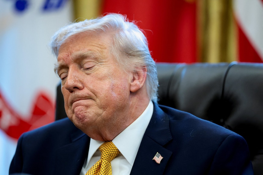A close up of Donald Trump with a grimace as he sits in the Oval Office in a suit and yellow tie