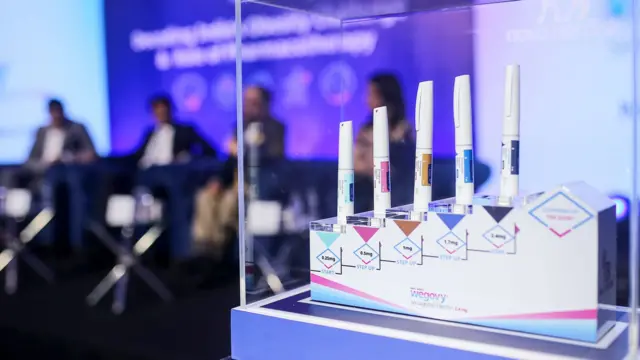 2025 年 6 月，在印度孟买举行的 “周纤达”（Wegovy）减肥药上市新闻发布会上，五支减肥药注射笔被放置在底座上的一个封闭支架里，台上坐着一些数人，他们坐在椅子上，面前是一块紫色的屏幕投影。