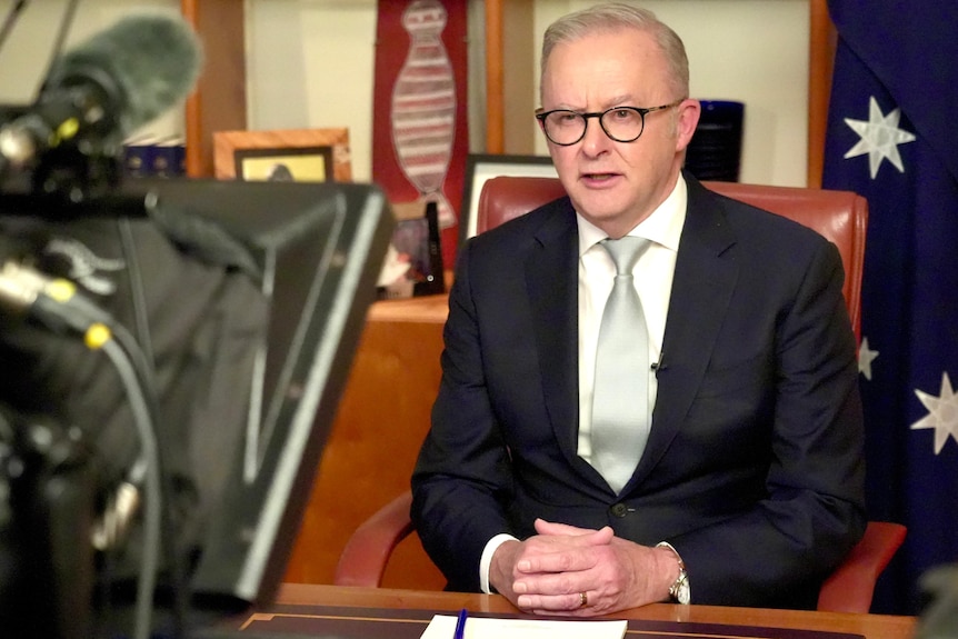 A man in a suit sits at a desk and reads a teleprompter, looking in to a camera.