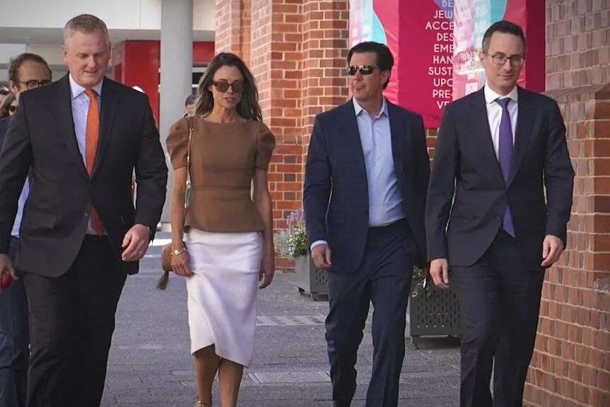 Bianca wears a brown top and white skirt as she walks next to John who wears a blue suit
