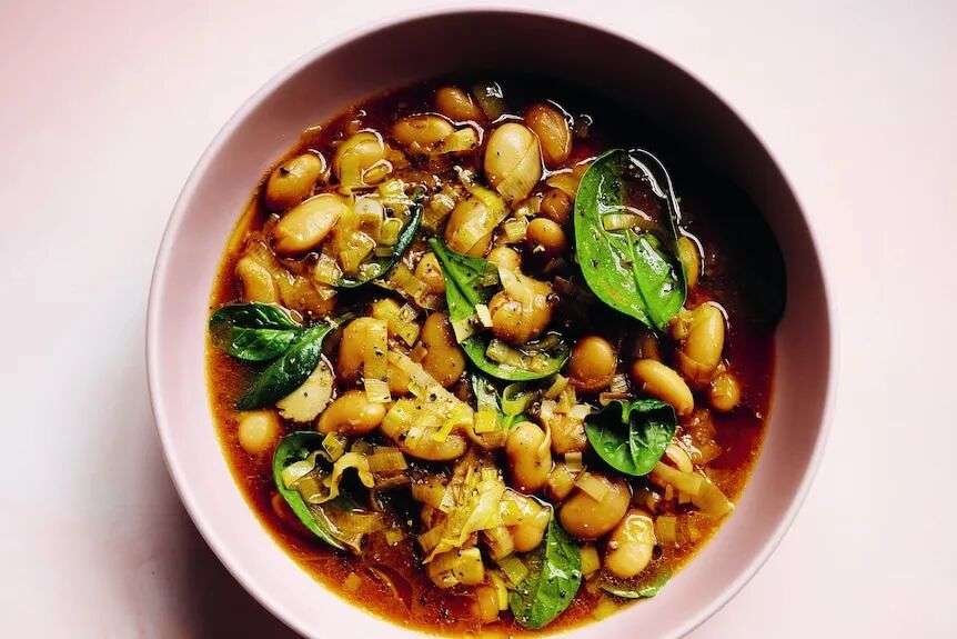 A pink bowl of brothy beans with visible spinach, against a pink background.