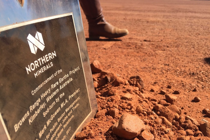 A sign explaining a new mine sits in red dirt as a person walks past in boots