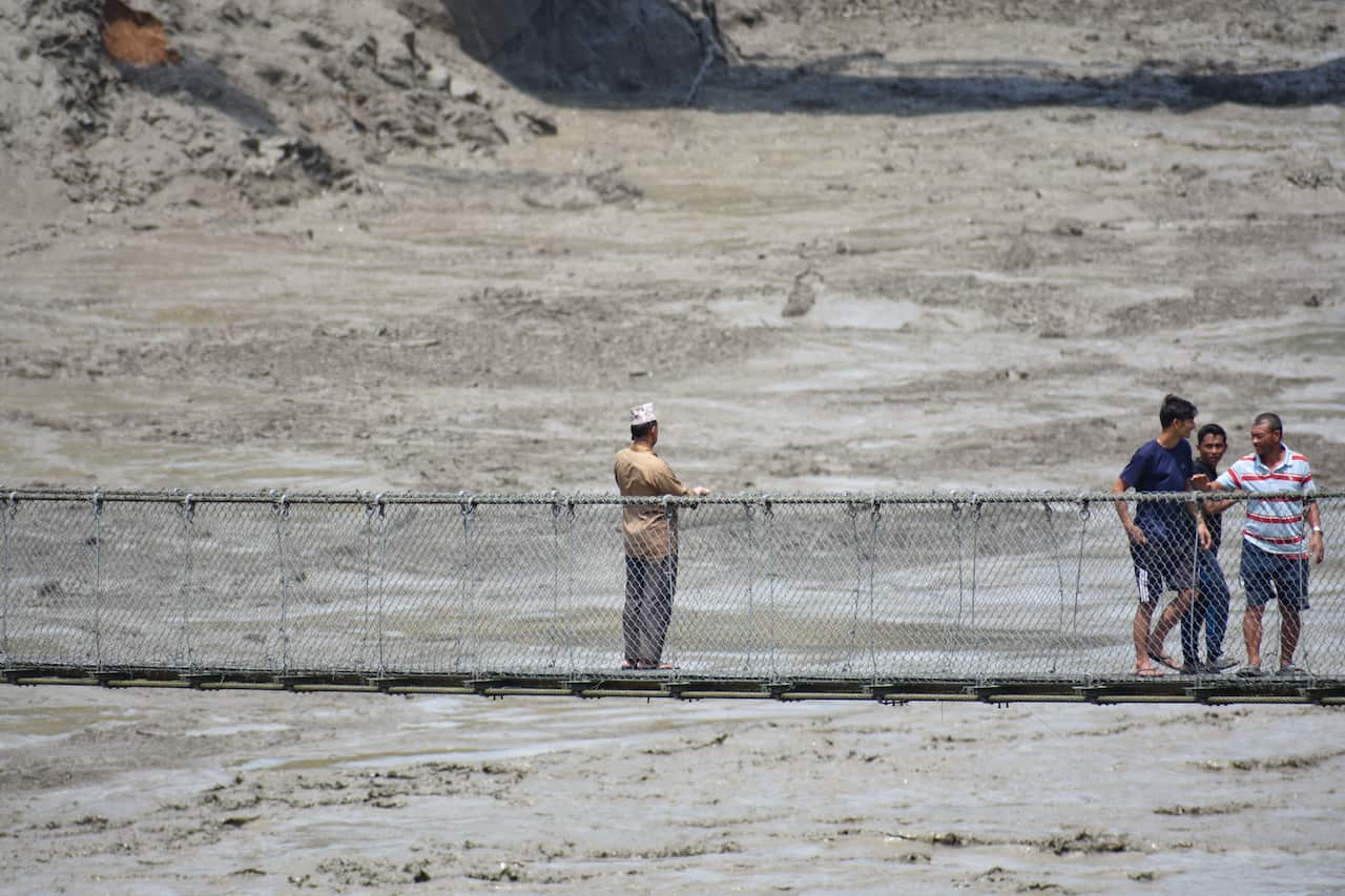 Several people standing on a suspension footbridge above a vast mud-covered landscape, with thick grey sediment stretching across the riverbed below.